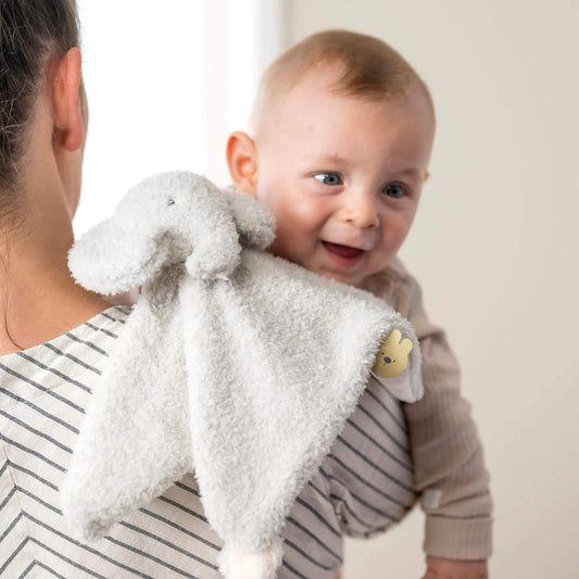 Smiling baby with an elephant doudou comfort blanket, showing soft grey plush material and soothing design features.
