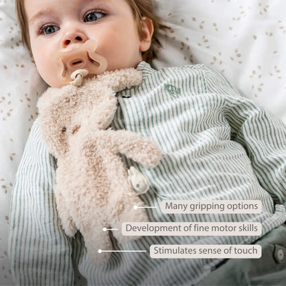 Baby holding a plush bunny pacifier holder against their chest, displayed with the text ‘Many gripping options’, ‘Development of fine motor skills’ and ‘Stimulates sense of touch’ on the image.