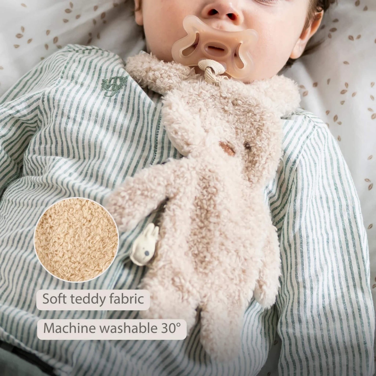 Plush bunny pacifier holder resting on a baby’s clothing with a close-up circle showing the teddy fabric texture, shown with the text ‘Soft teddy fabric’ and ‘Machine washable 30°’ on the image.