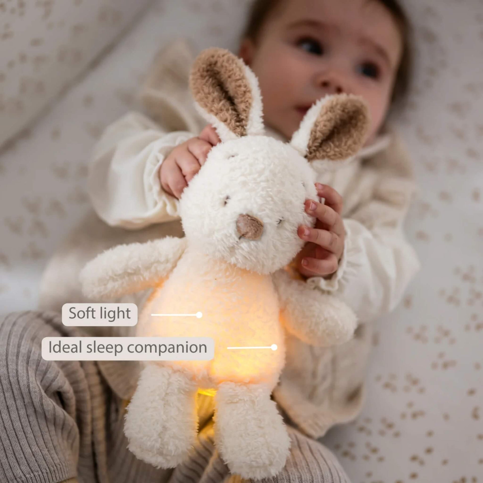 A baby lying on patterned bedding while holding a glowing plush bunny nightlight close to their chest.