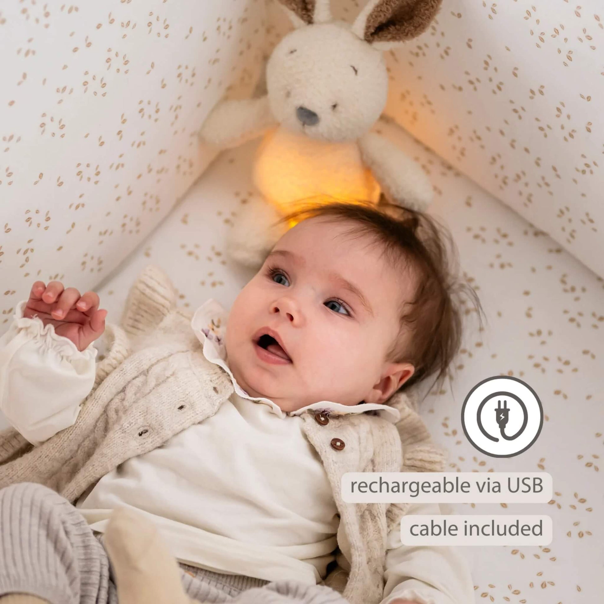 A baby lying in a cot with a glowing plush bunny nightlight positioned behind them, with a USB charging icon highlighted.