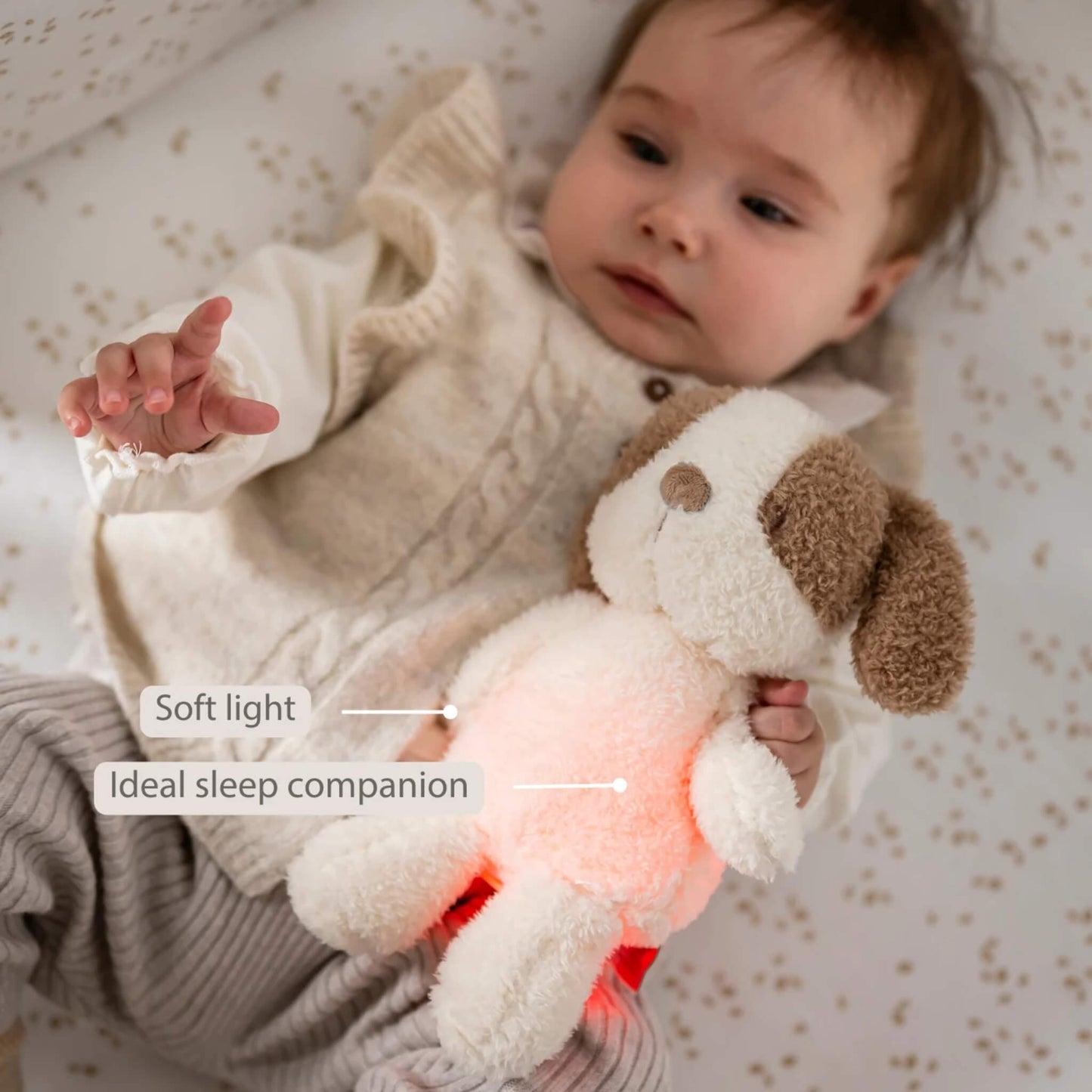 A baby lying on a patterned sheet while holding a glowing plush dog nightlight close to their chest.
