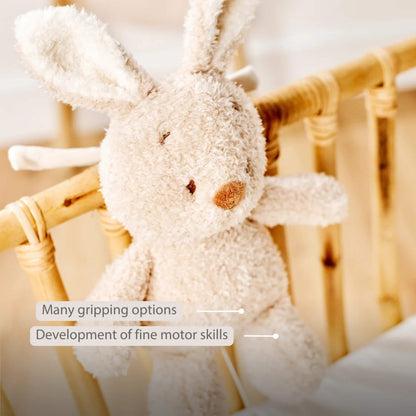 A plush bunny musical toy hanging from a wooden cot rail with a textured body and long ears. On-image text highlights “Many gripping options” and “Development of fine motor skills”.