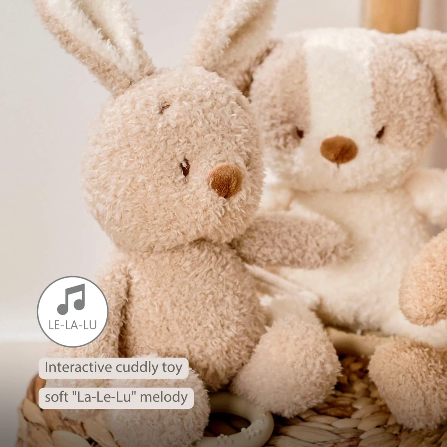 A plush bunny toy sitting in a woven basket beside another soft toy, with the pull-cord ring partly visible. On-image text reads “Interactive cuddly toy” and “Soft ‘La-Le-Lu’ melody”.