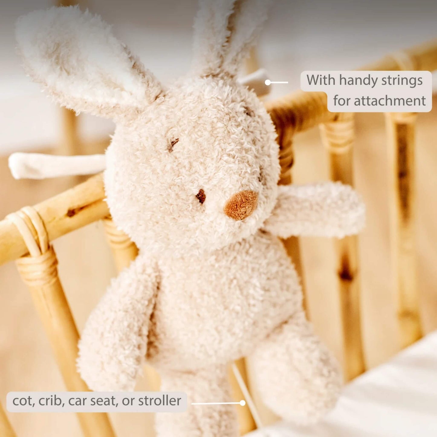 A soft plush bunny tied to a wooden cot rail with its pull-cord hanging down. On-image text states “With handy strings for attachment” and lists “cot, crib, car seat or stroller”.