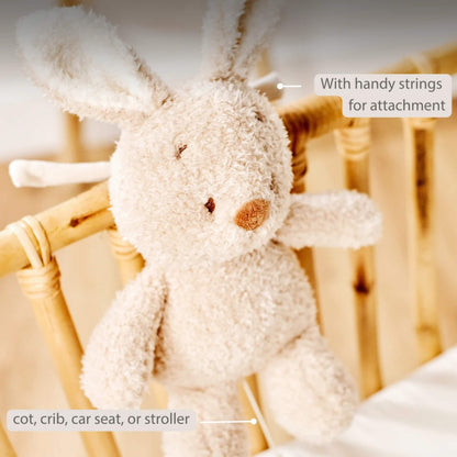 A soft plush bunny tied to a wooden cot rail with its pull-cord hanging down. On-image text states “With handy strings for attachment” and lists “cot, crib, car seat or stroller”.