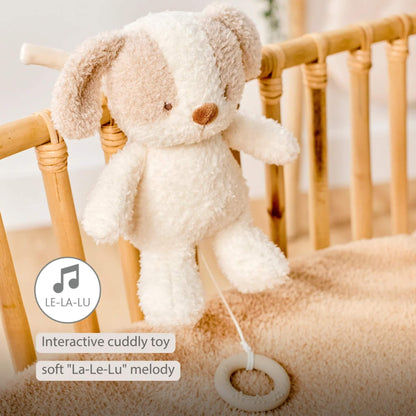 A plush dog musical toy hangs from a cot rail with the silicone pull ring visible, shown with on-image text stating “Interactive cuddly toy” and “soft ‘La-Le-Lu’ melody.”