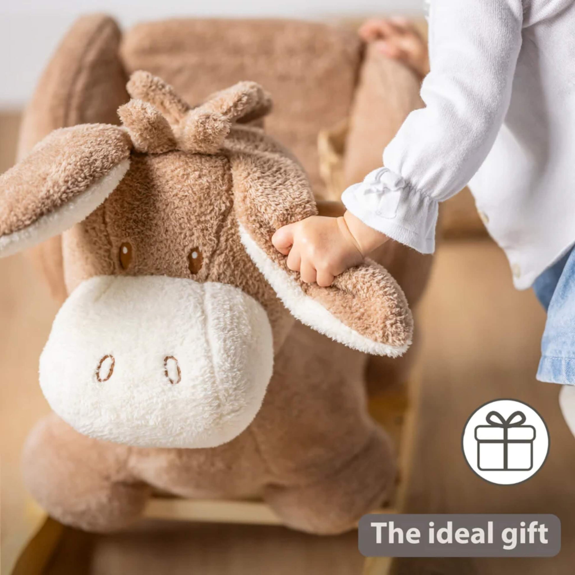 Close-up of a child’s hand gripping a soft plush donkey ear while sitting on a rocking toy in a warm home interior.