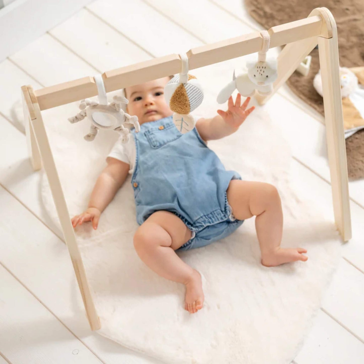 Baby lying on a soft play mat reaching up toward hanging sensory toys on a wooden play arch, encouraging grasping and early coordination.