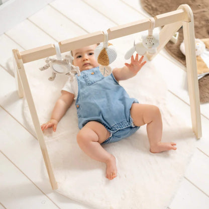 Baby lying on a soft play mat reaching up toward hanging sensory toys on a wooden play arch, encouraging grasping and early coordination.