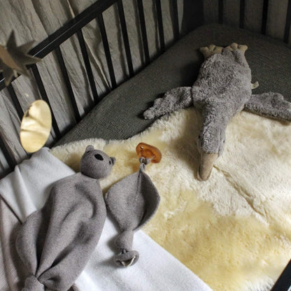 Cream sheepskin baby rug placed inside a dark cot with soft grey and white blankets, a pacifier, and two plush toys resting on top.