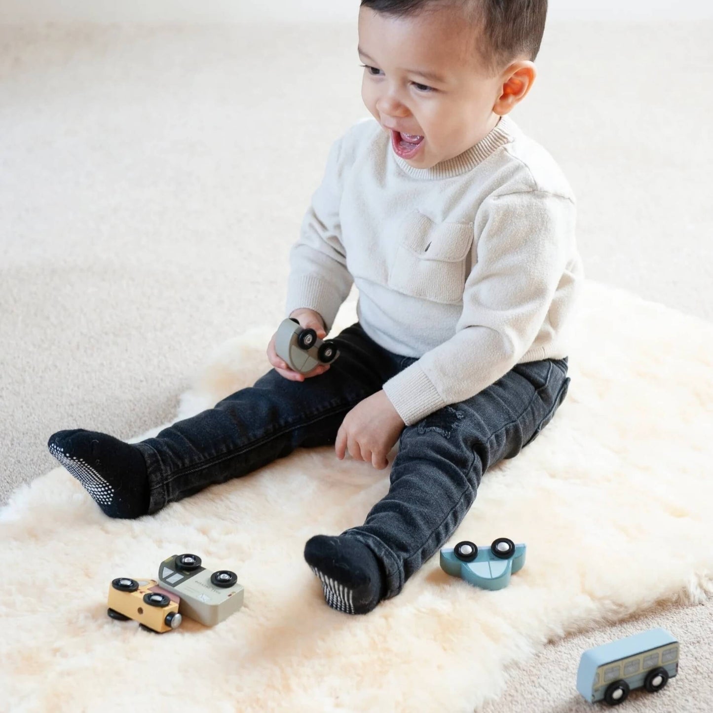 Smiling toddler sits on a sheepskin baby rug on the floor, holding wooden toy cars with more toys scattered nearby.