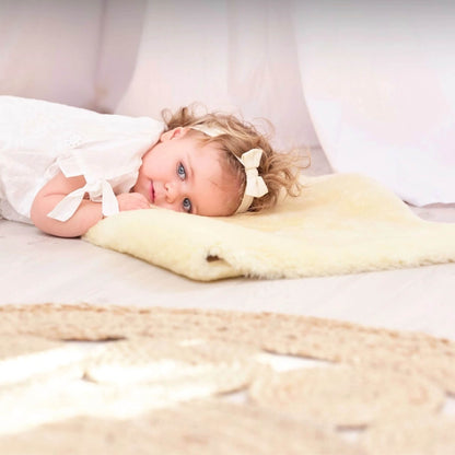 Young girl lies resting on a cream sheepskin baby rug indoors, wearing a white dress with a bow headband, gazing softly at the camera.