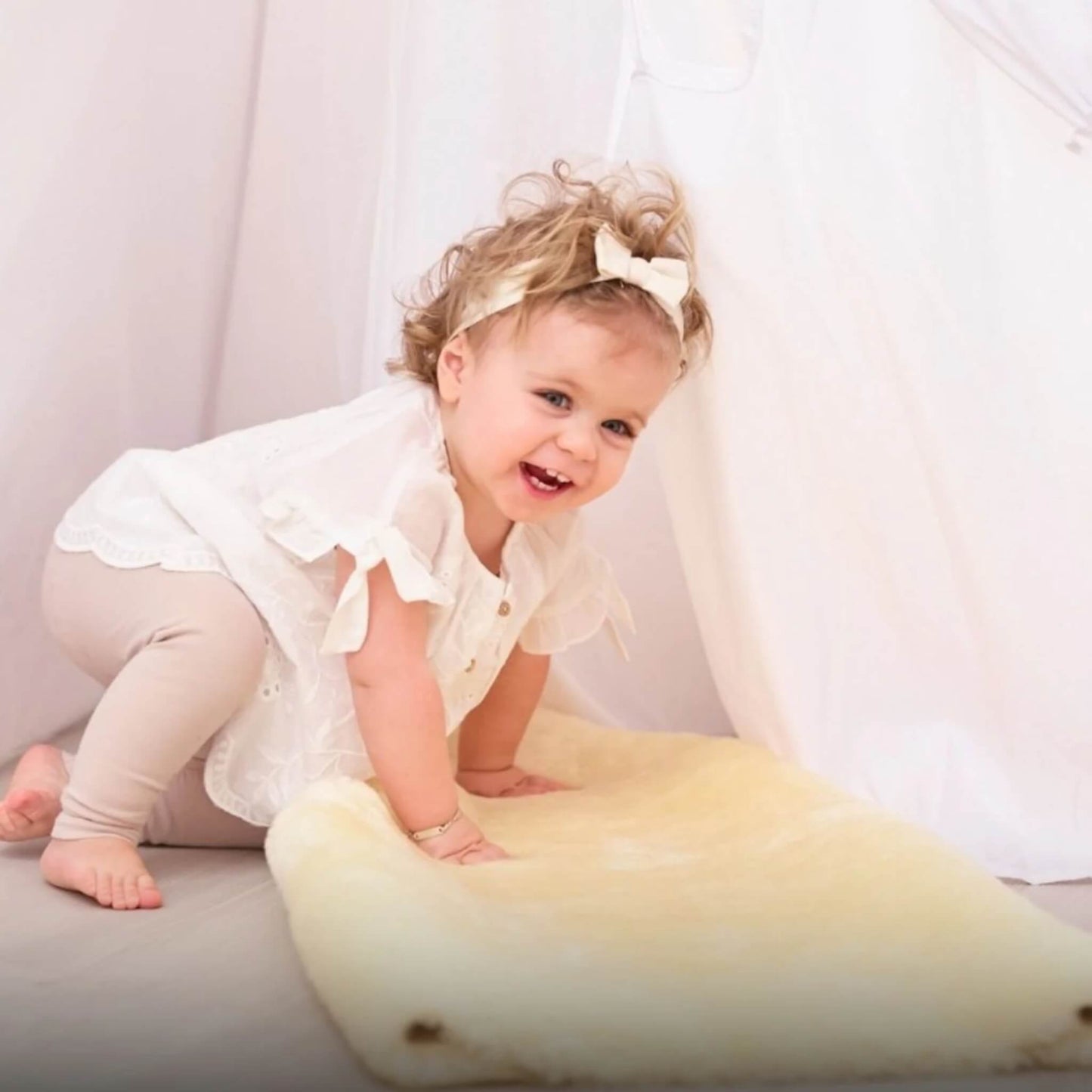 Toddler in a white top smiles while kneeling on a cream sheepskin rug, leaning forward in play.