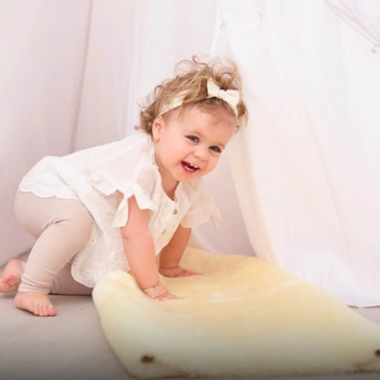 Toddler in a white top smiles while kneeling on a cream sheepskin rug, leaning forward in play.