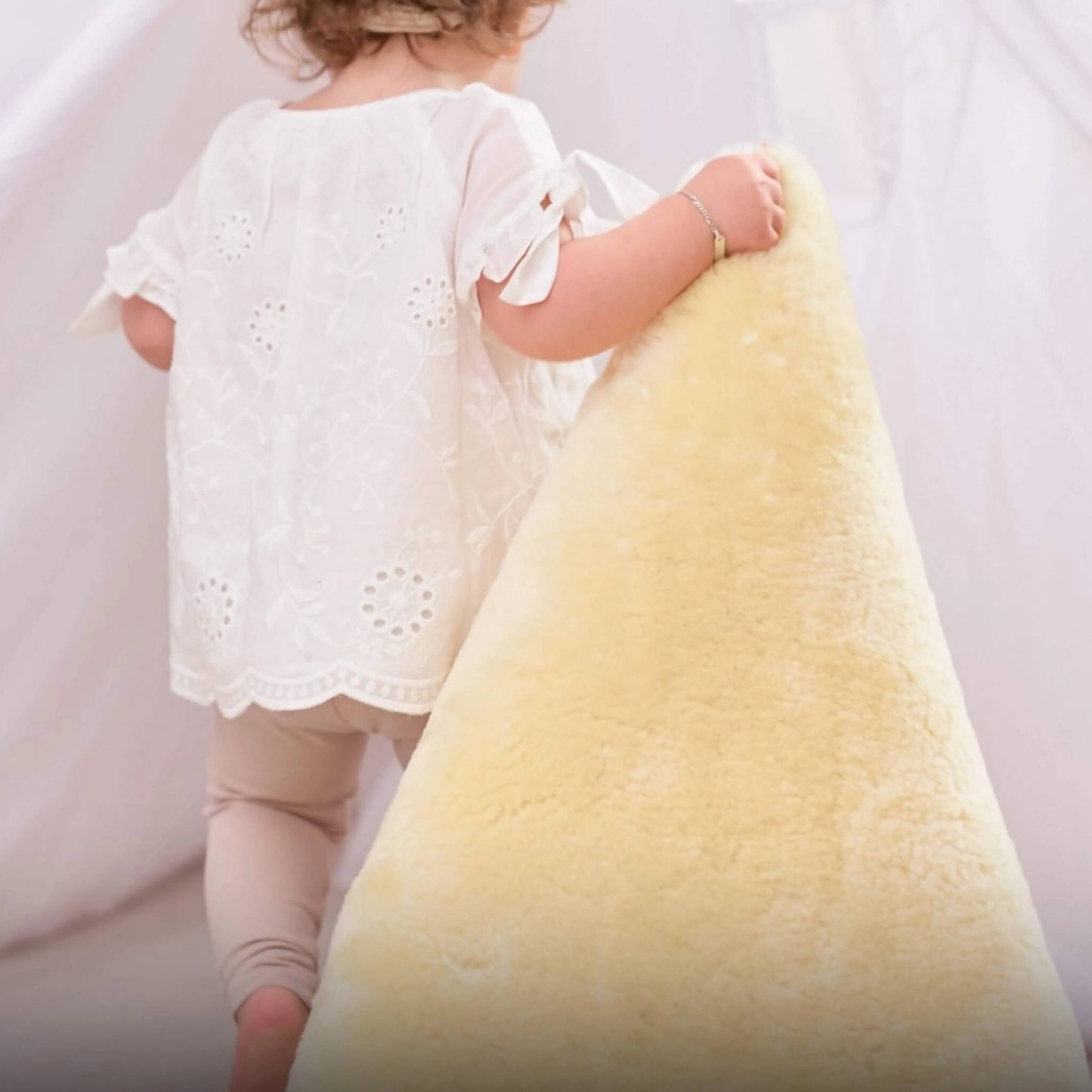 Toddler lifts and carries the edge of a cream sheepskin rug while walking indoors, showing the rug’s thickness and softness.