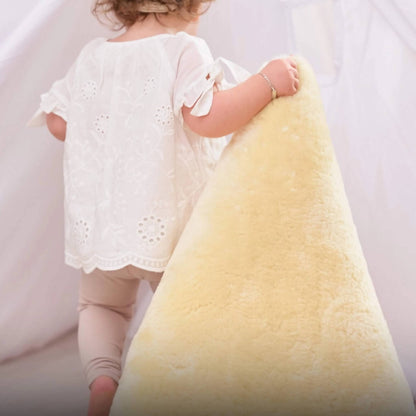 Toddler lifts and carries the edge of a cream sheepskin rug while walking indoors, showing the rug’s thickness and softness.