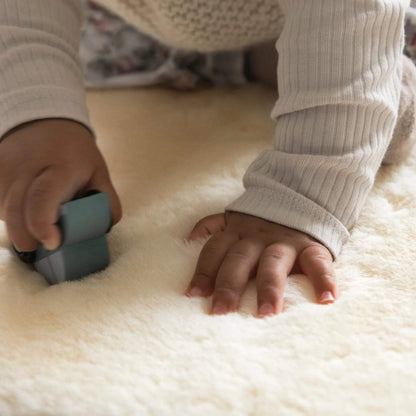 Close-up of a child’s hand pressing onto a cream sheepskin rug while playing with a blue wooden toy vehicle.