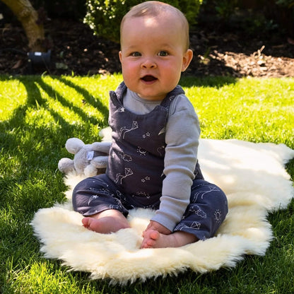 Baby sits outdoors on a golden sheepskin rug spread on grass, dressed in grey dungarees and holding a soft toy rabbit.