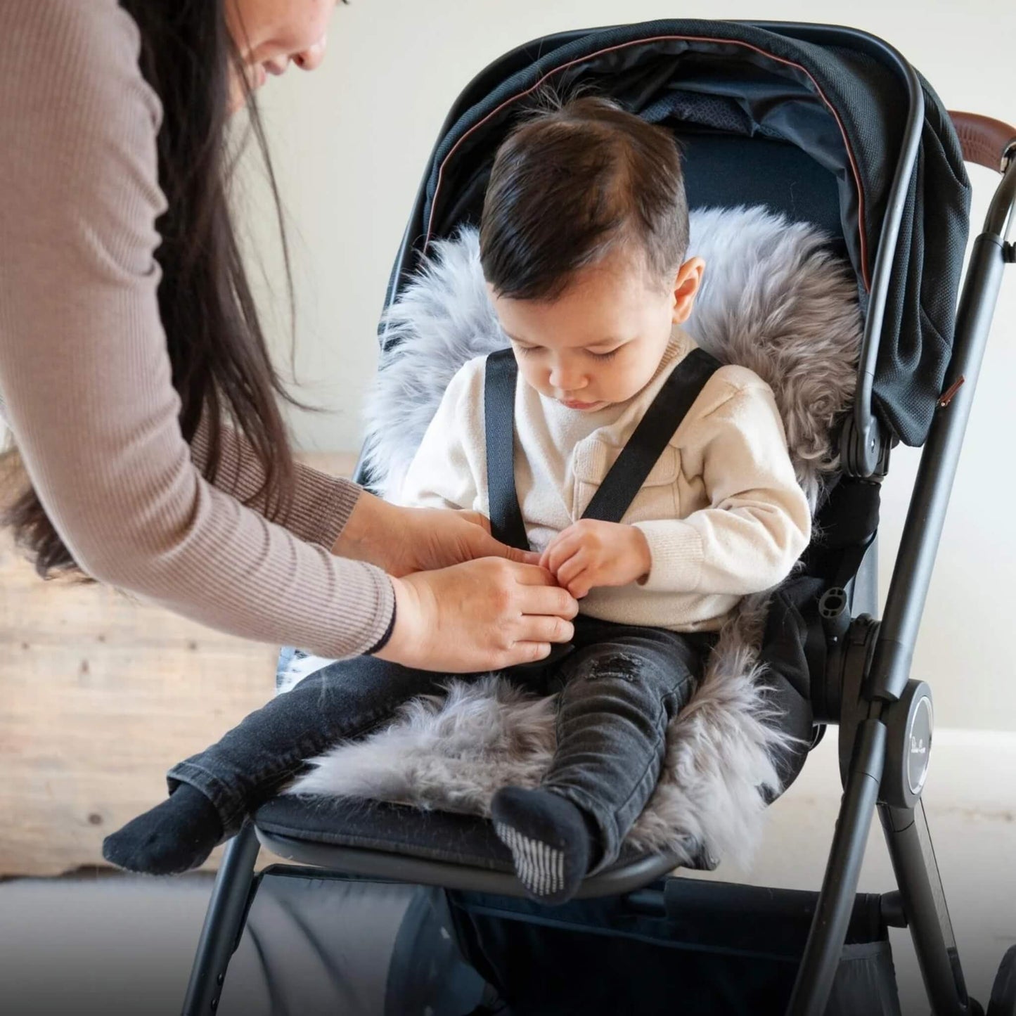 Toddler seated in a pushchair lined with grey sheepskin pram liner while mother secures harness straps.