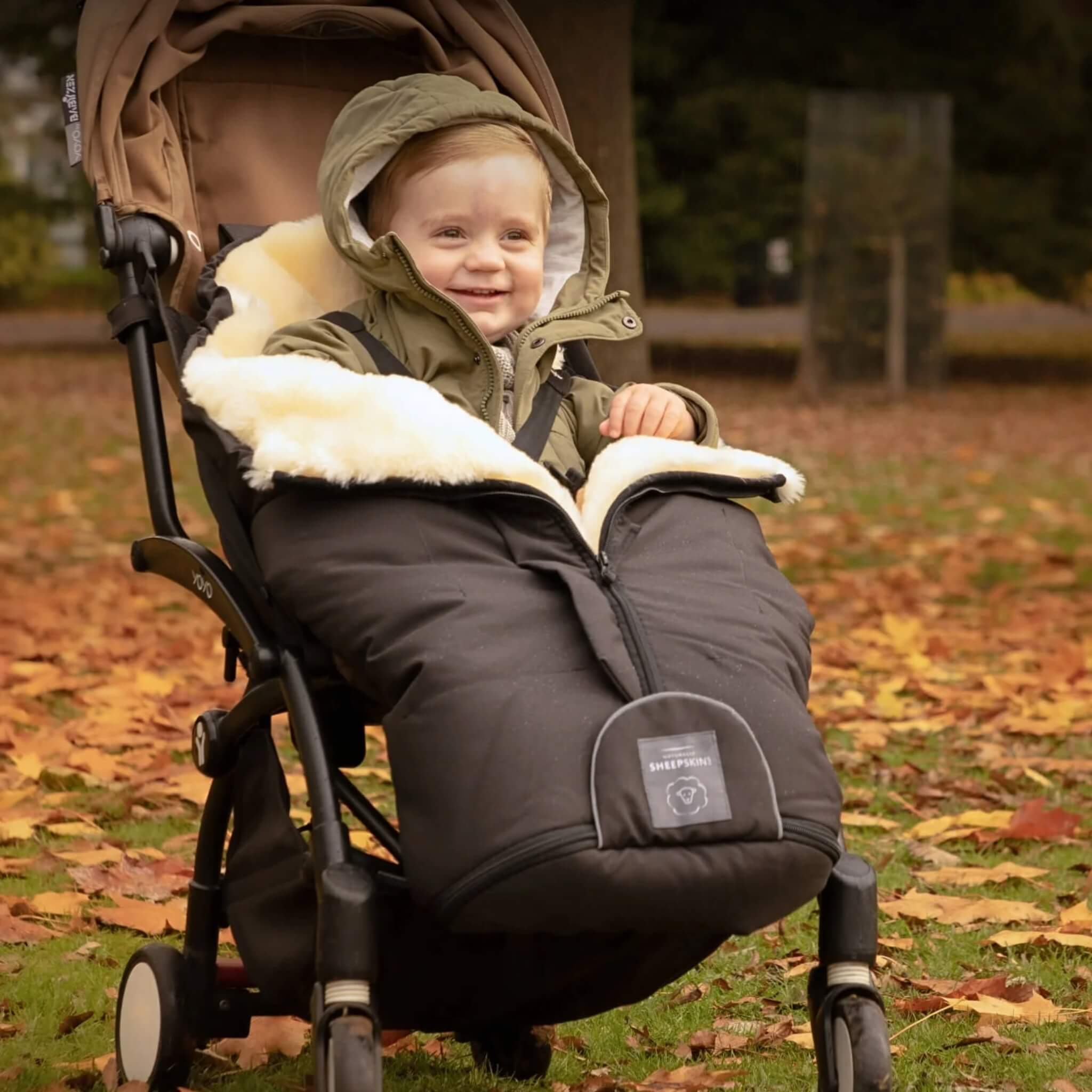 Toddler in a pushchair outdoors in autumn, smiling while wrapped in a black footmuff with cream sheepskin interior.