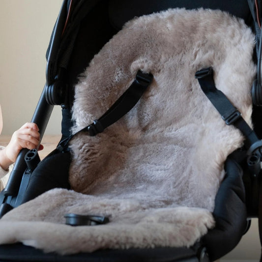 Close-up view of a taupe sheepskin pram liner fitted in a stroller, showing the texture of the wool and harness straps resting on top.