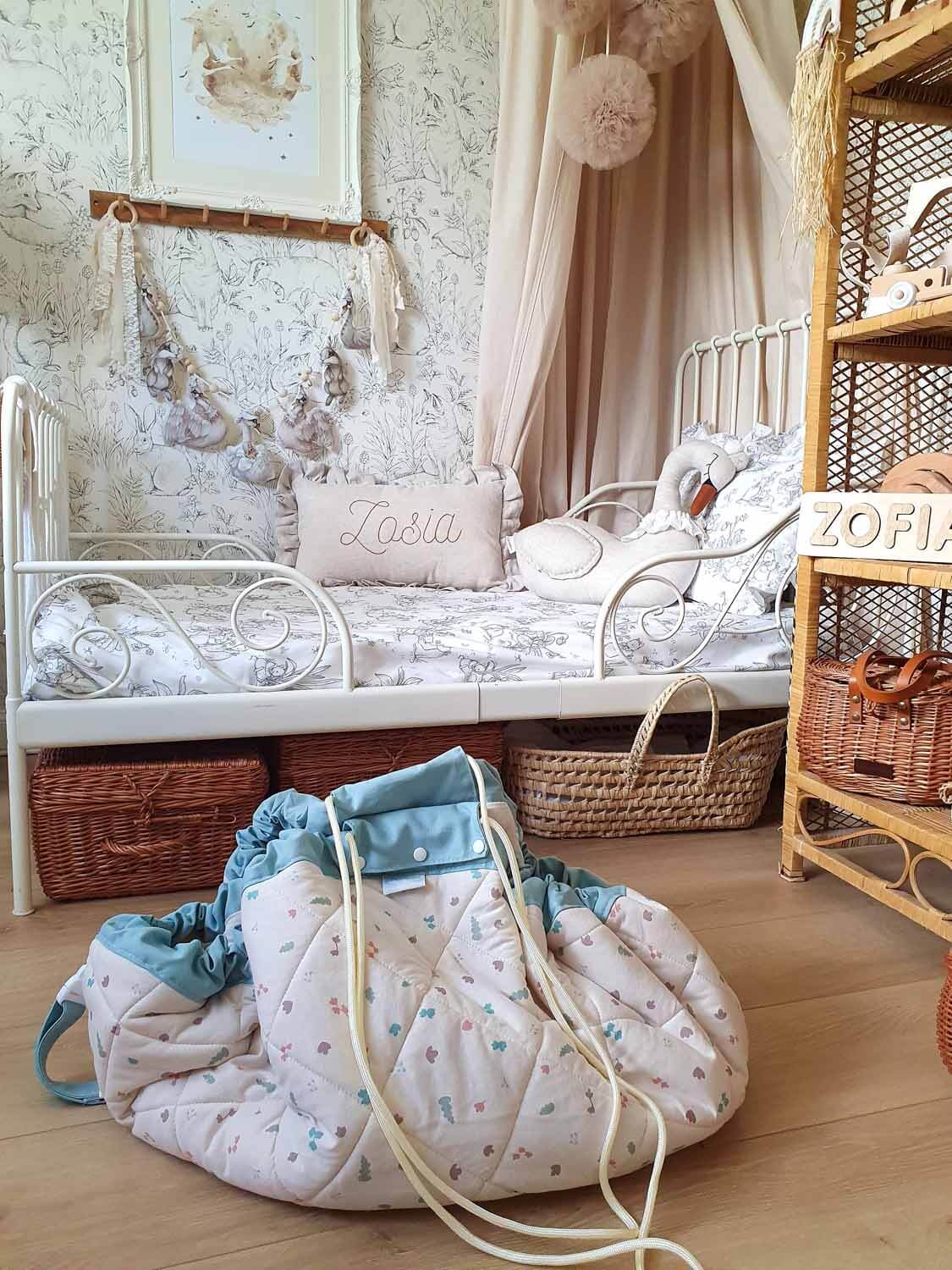 A styled children’s bedroom showing a gathered toy storage bag resting on the floor beside a daybed and soft décor.
