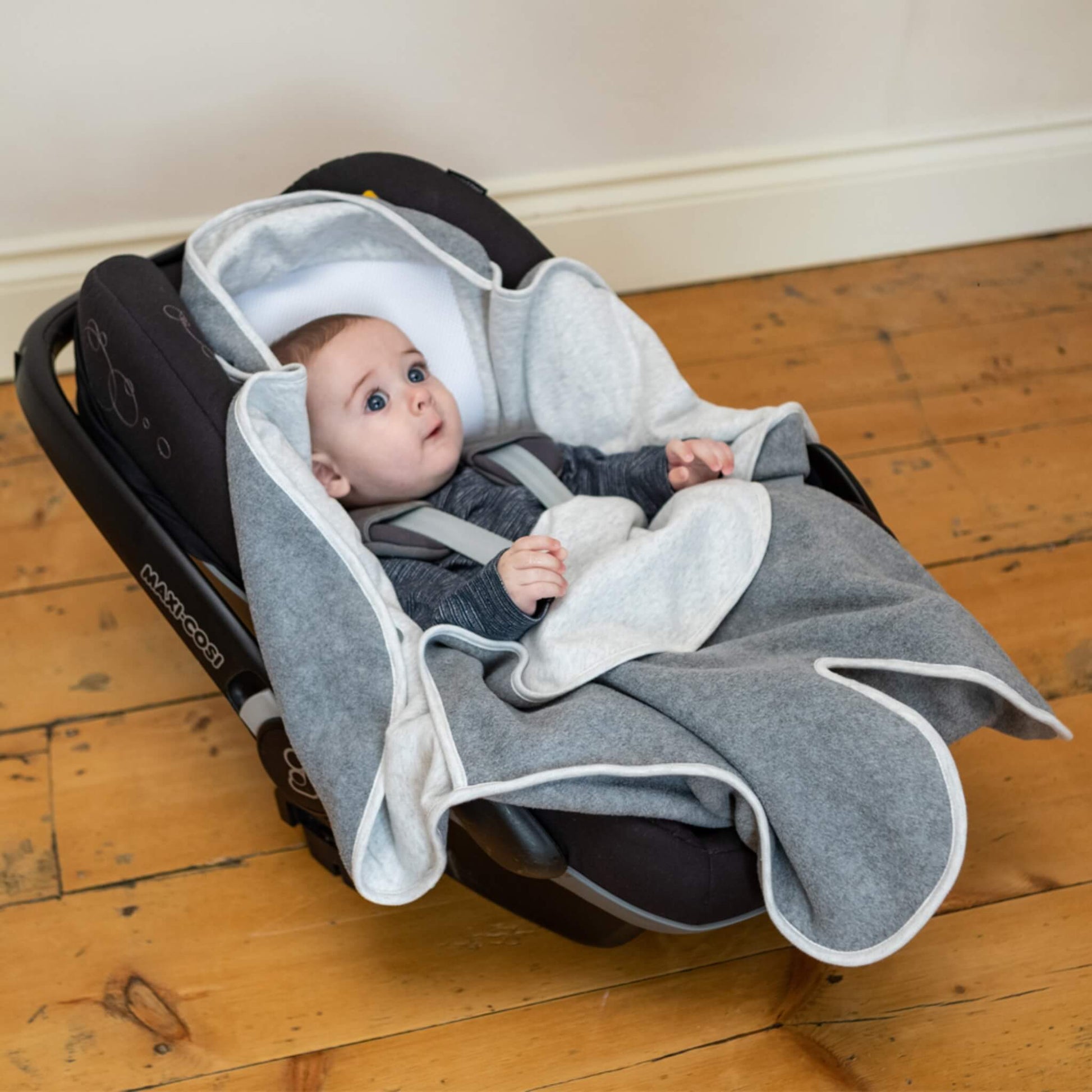 Baby seated in a car seat with light grey and darker grey blanket panels arranged loosely around the body.