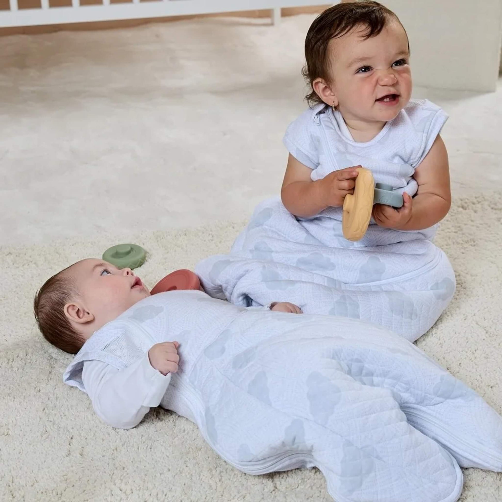 Two babies wearing quilted sleep bags with cloud patterns, one lying on a cream rug and the other sitting up holding toys.