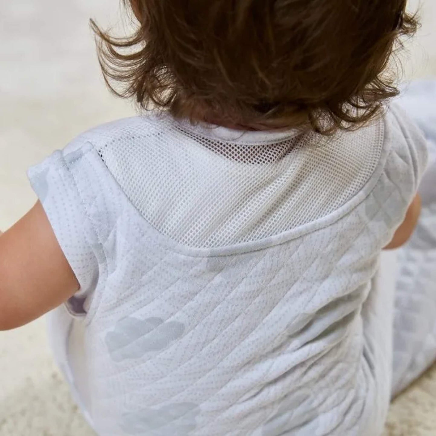 A back view of a baby sitting while wearing a sleeveless quilted sleep bag, showing a breathable mesh panel across the shoulders.
