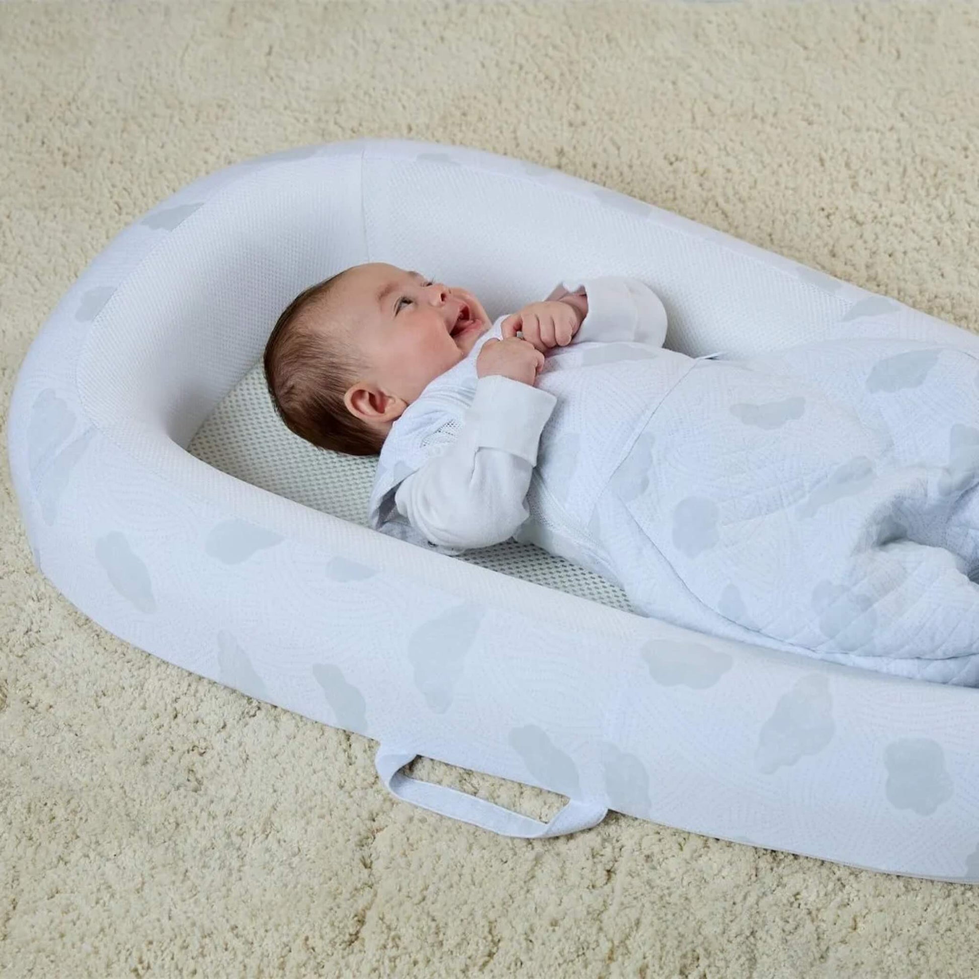 A baby bed placed on a carpeted floor, showing the carry handle stitched into the side panel.