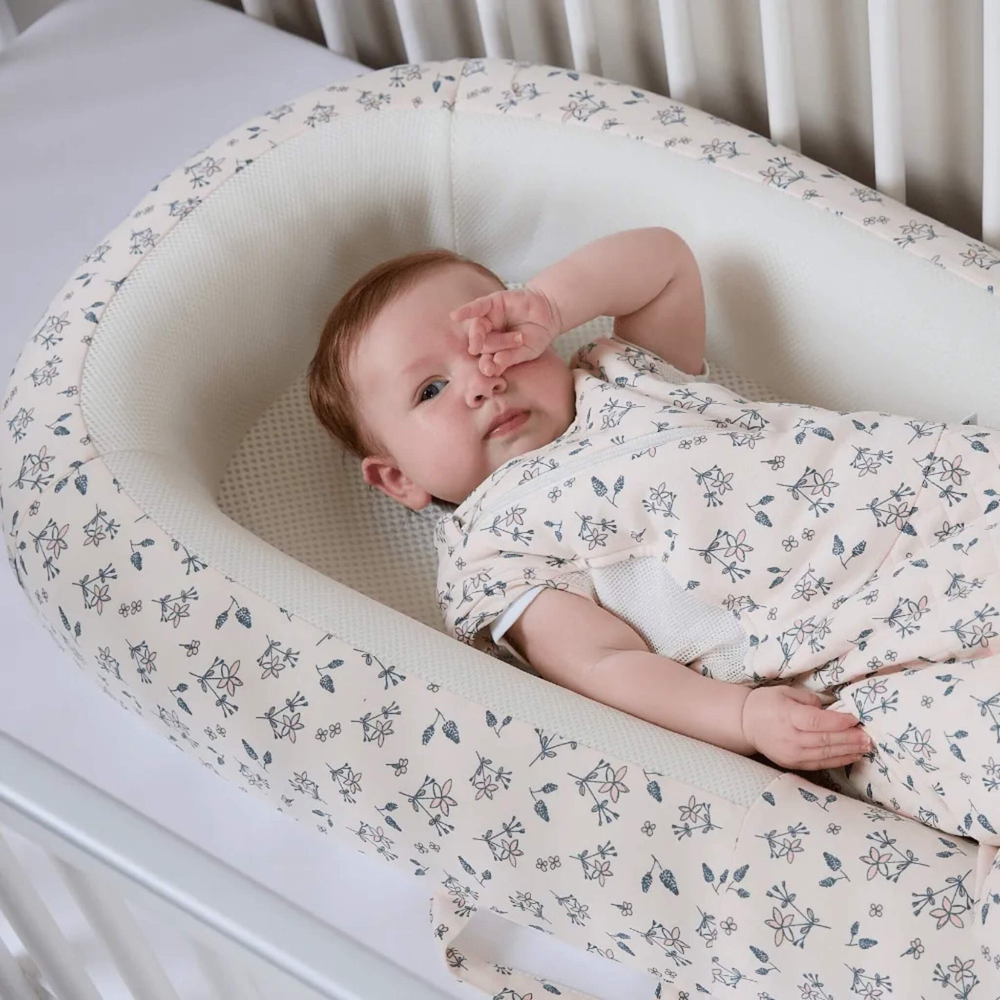 A side view of a baby bed showing raised, structured sides with a soft floral fabric finish.