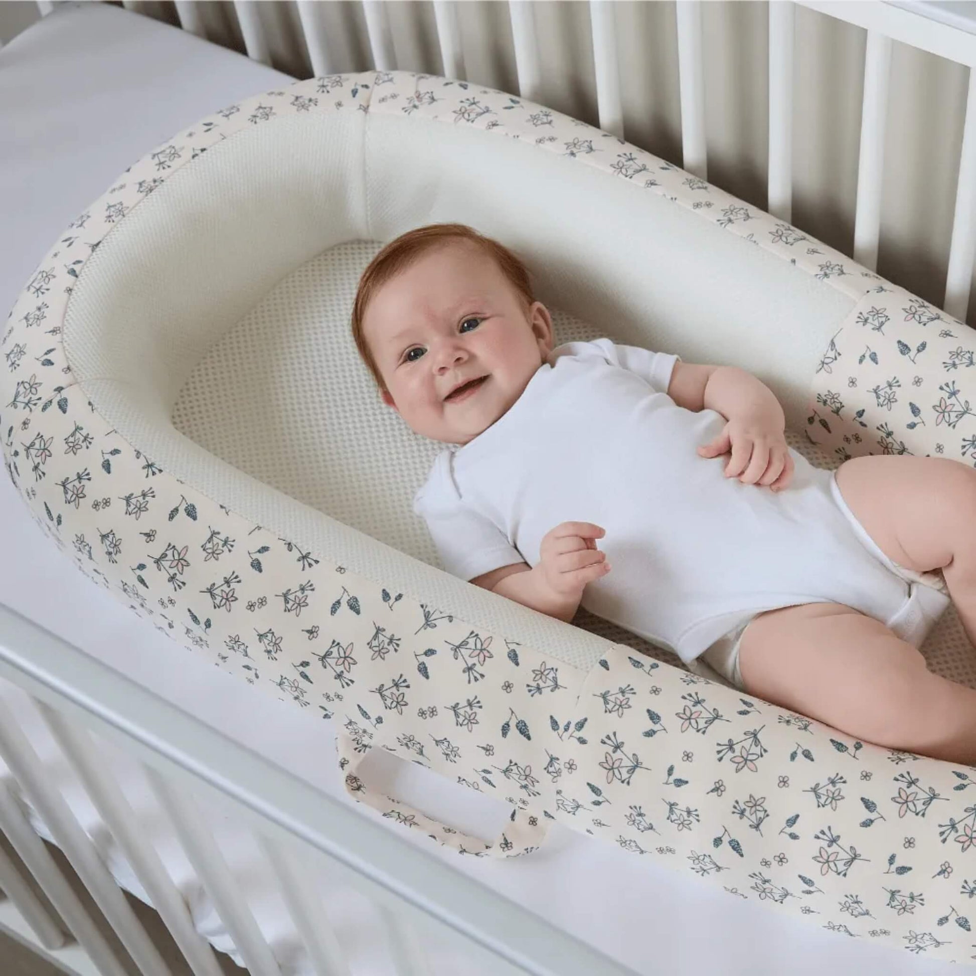 A baby lying on a firm, flat mesh mattress inside a structured baby bed placed in a cot.