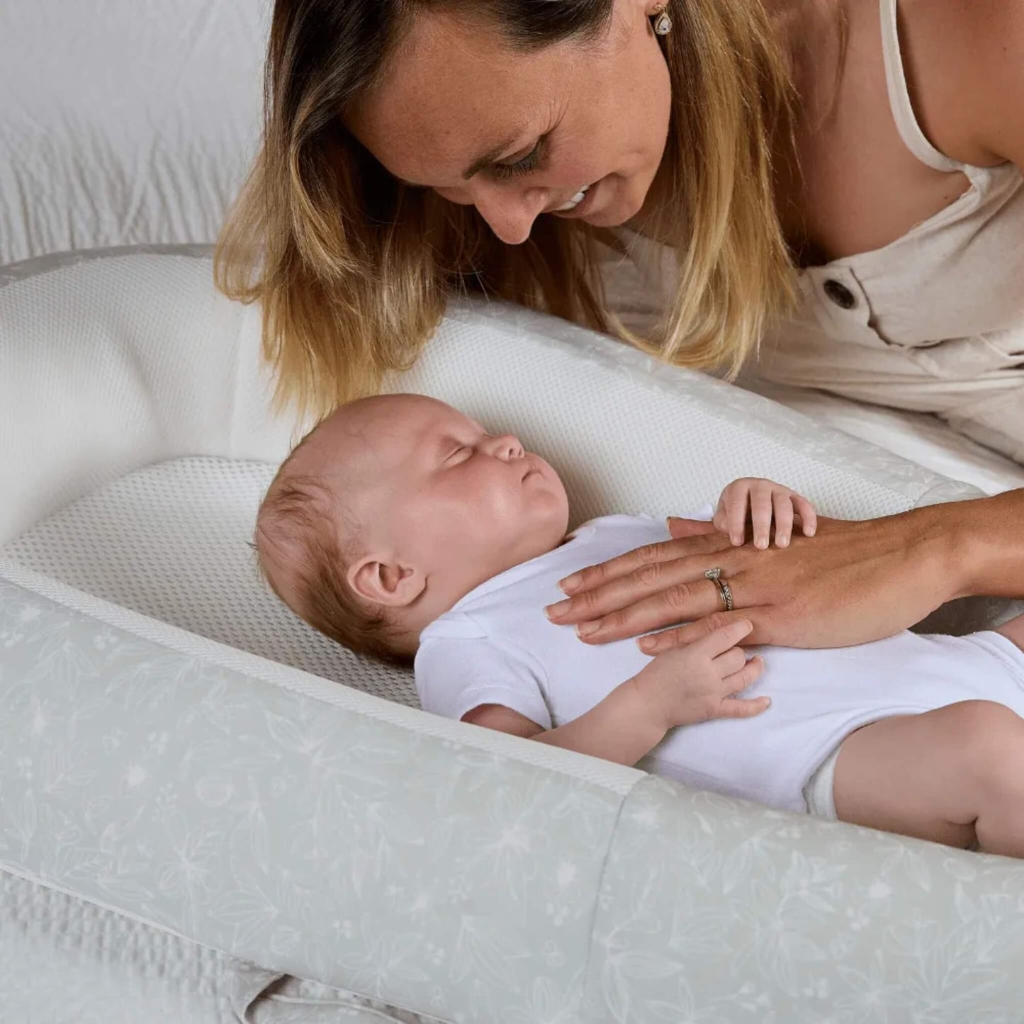 A mother leaning over a baby resting in a baby bed, showing the baby supported by firm sides and a breathable mesh base.