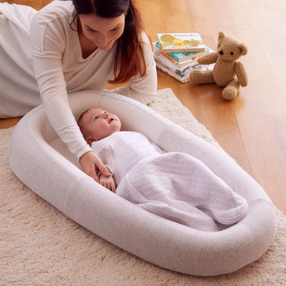 A baby resting inside a structured baby bed with mesh base, placed on a rug beside nursery furniture in a calm home setting.