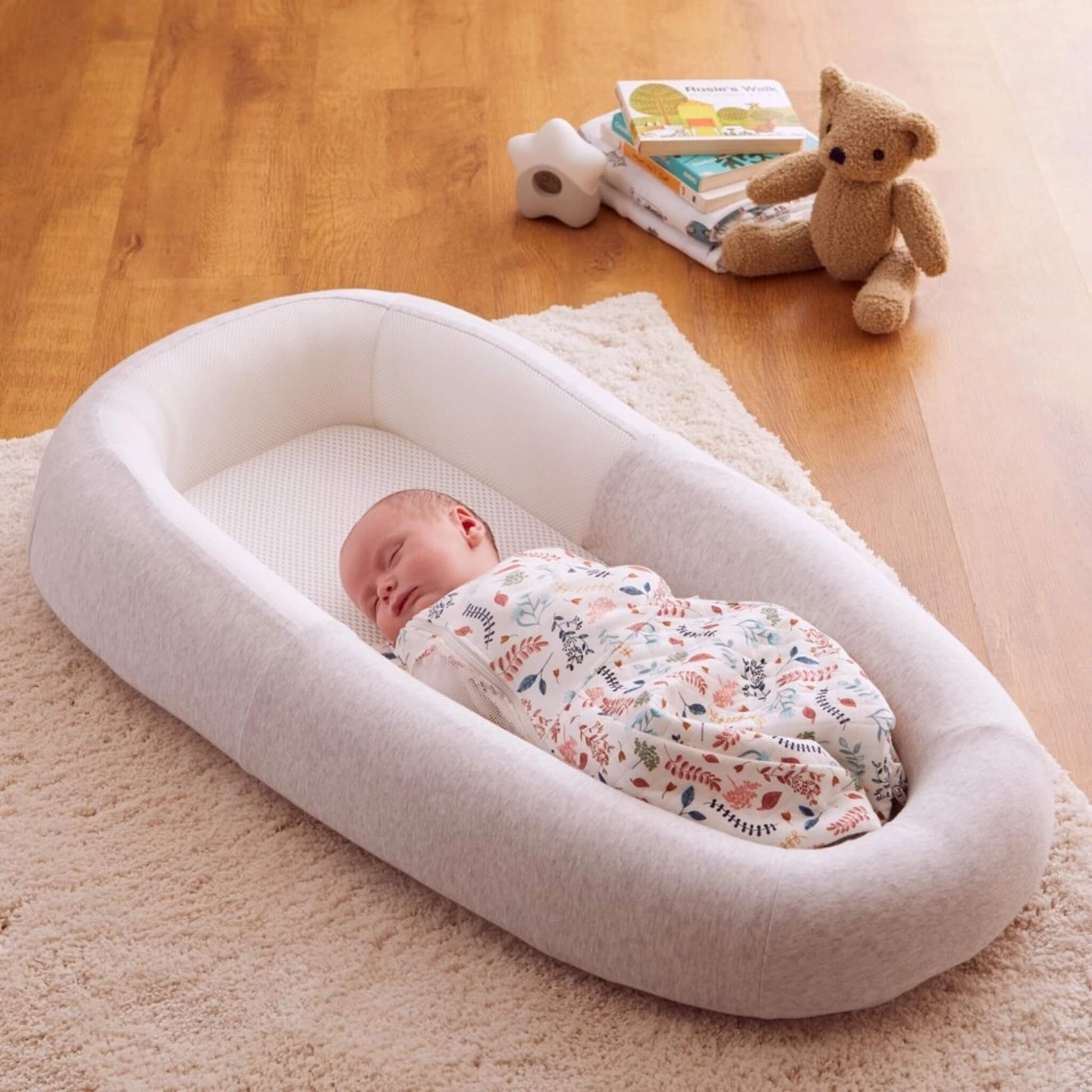 A baby lying comfortably inside a grey baby bed with breathable mesh panels, positioned on a soft rug during daytime rest.