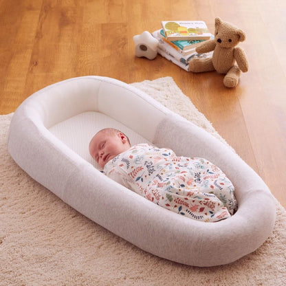 A baby lying comfortably inside a grey baby bed with breathable mesh panels, positioned on a soft rug during daytime rest.
