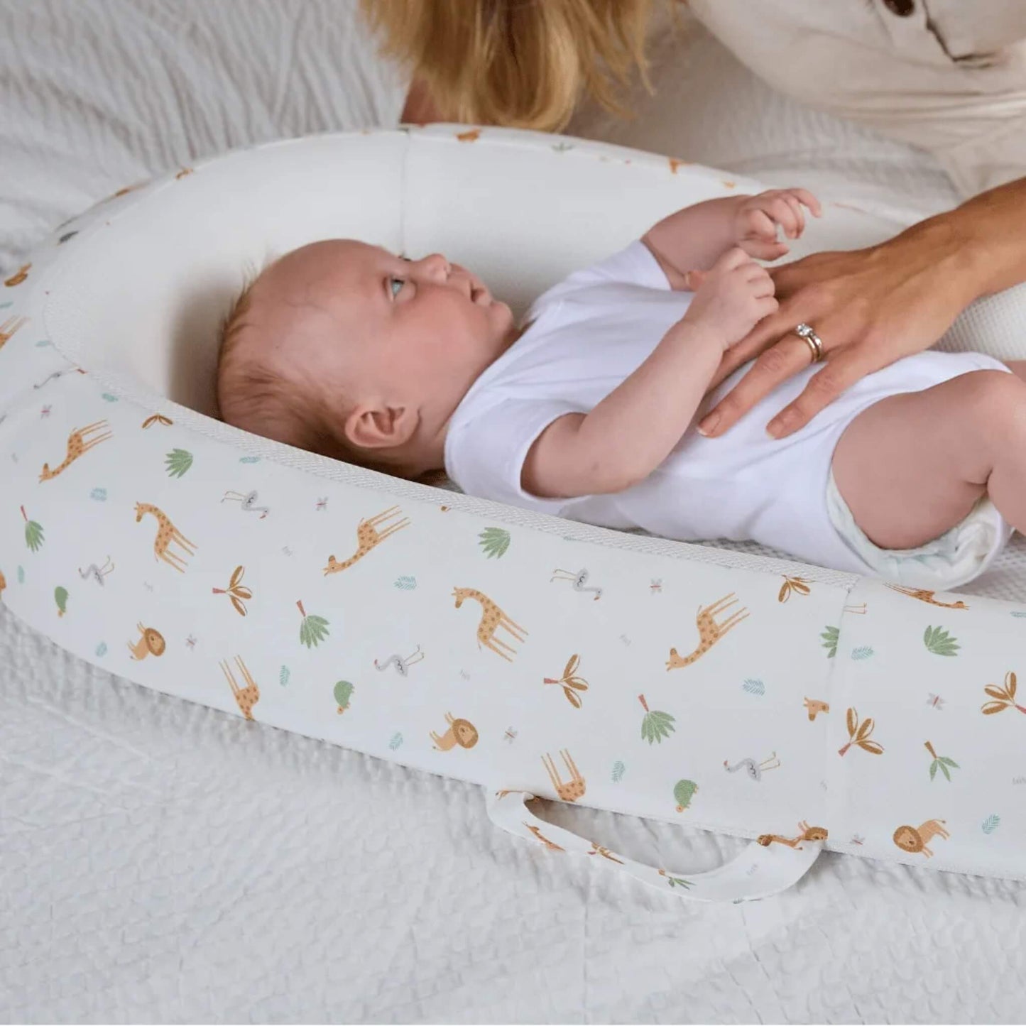 A baby lying on a breathable mesh mattress inside a structured baby bed, with an adult hand resting gently on the baby’s torso.