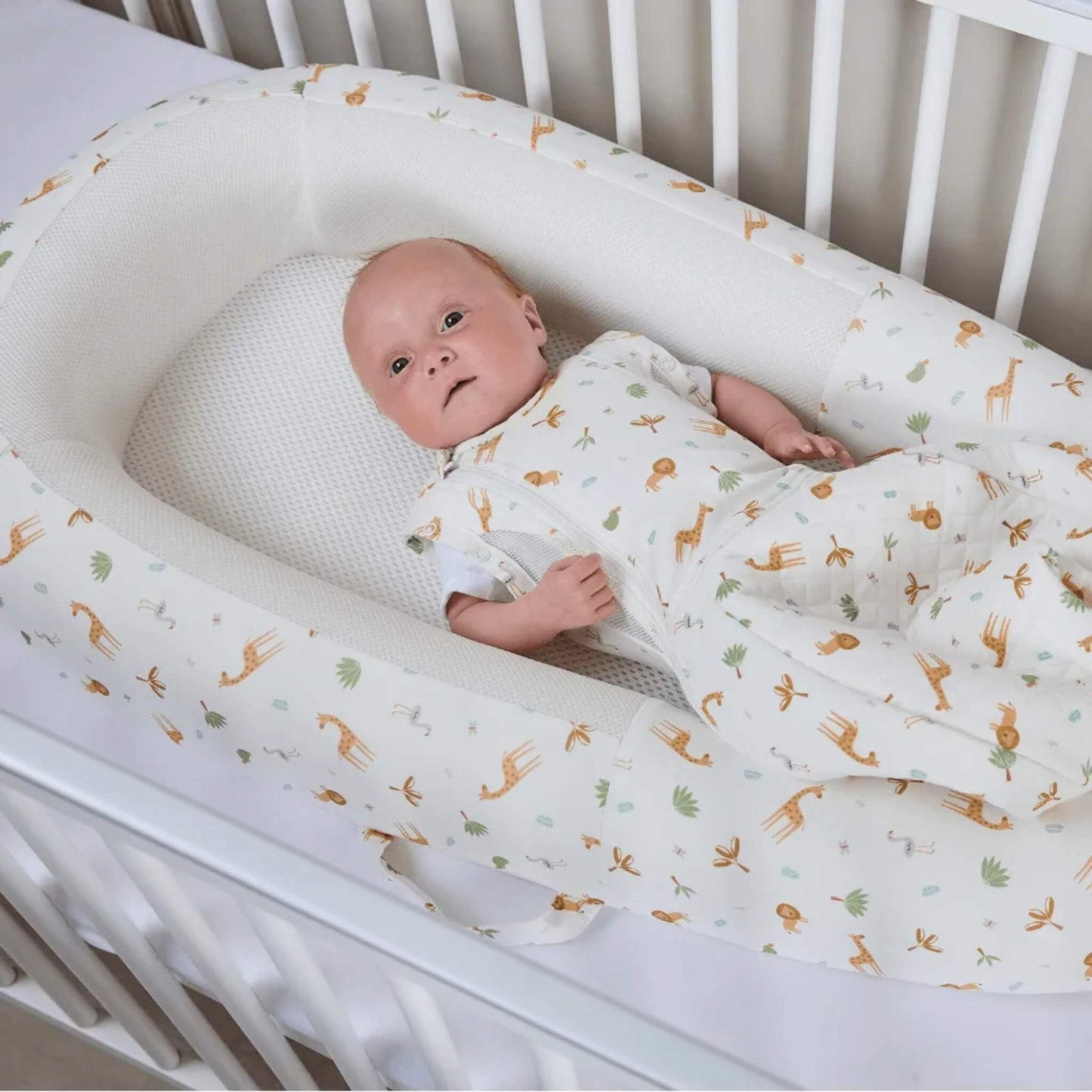 A baby resting inside a baby bed placed on a cot mattress, showing the depth of the sides and the flat mesh sleep surface.