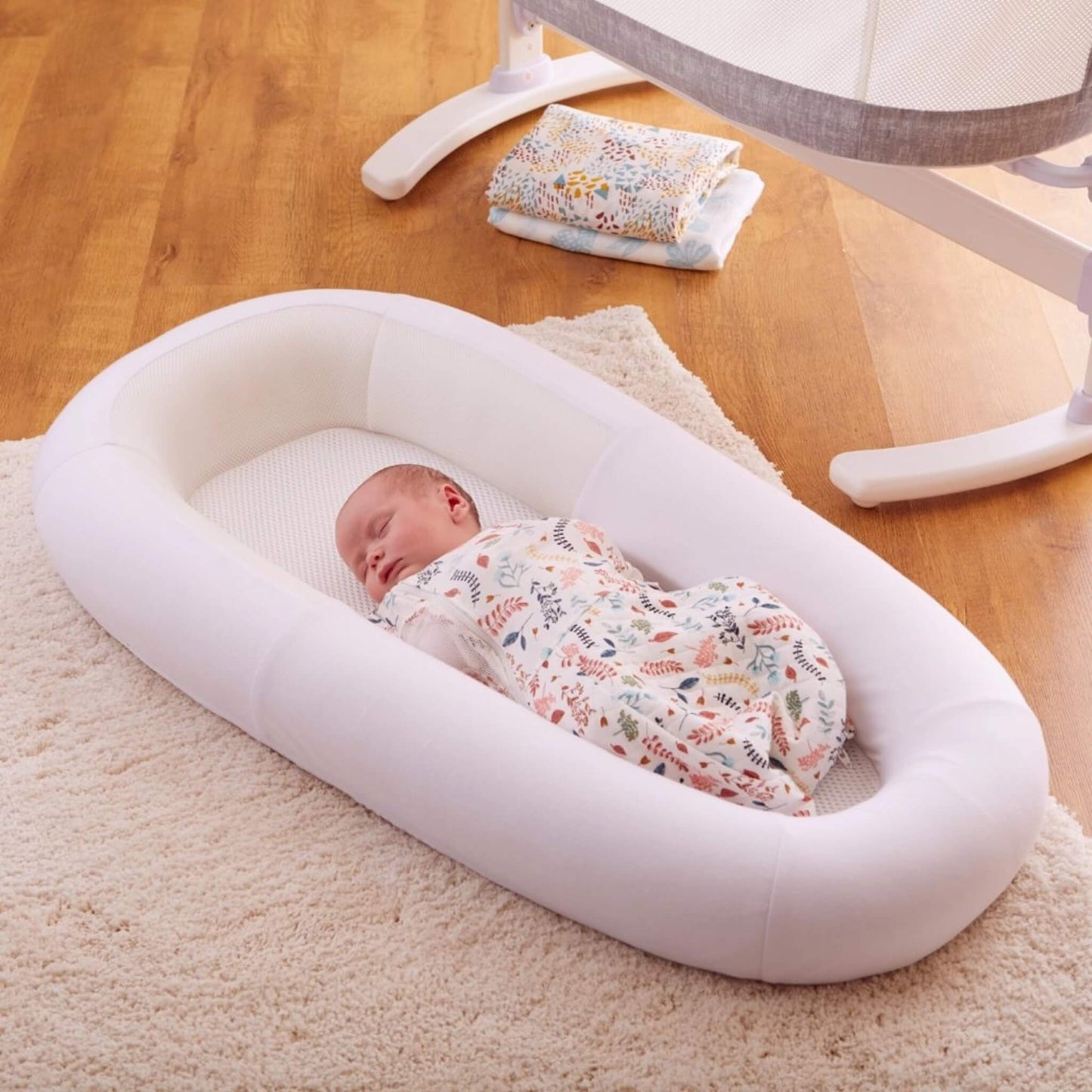 A baby resting on a mesh mattress inside a structured baby bed, placed on a rug beside nursery furniture in a calm daytime setting.