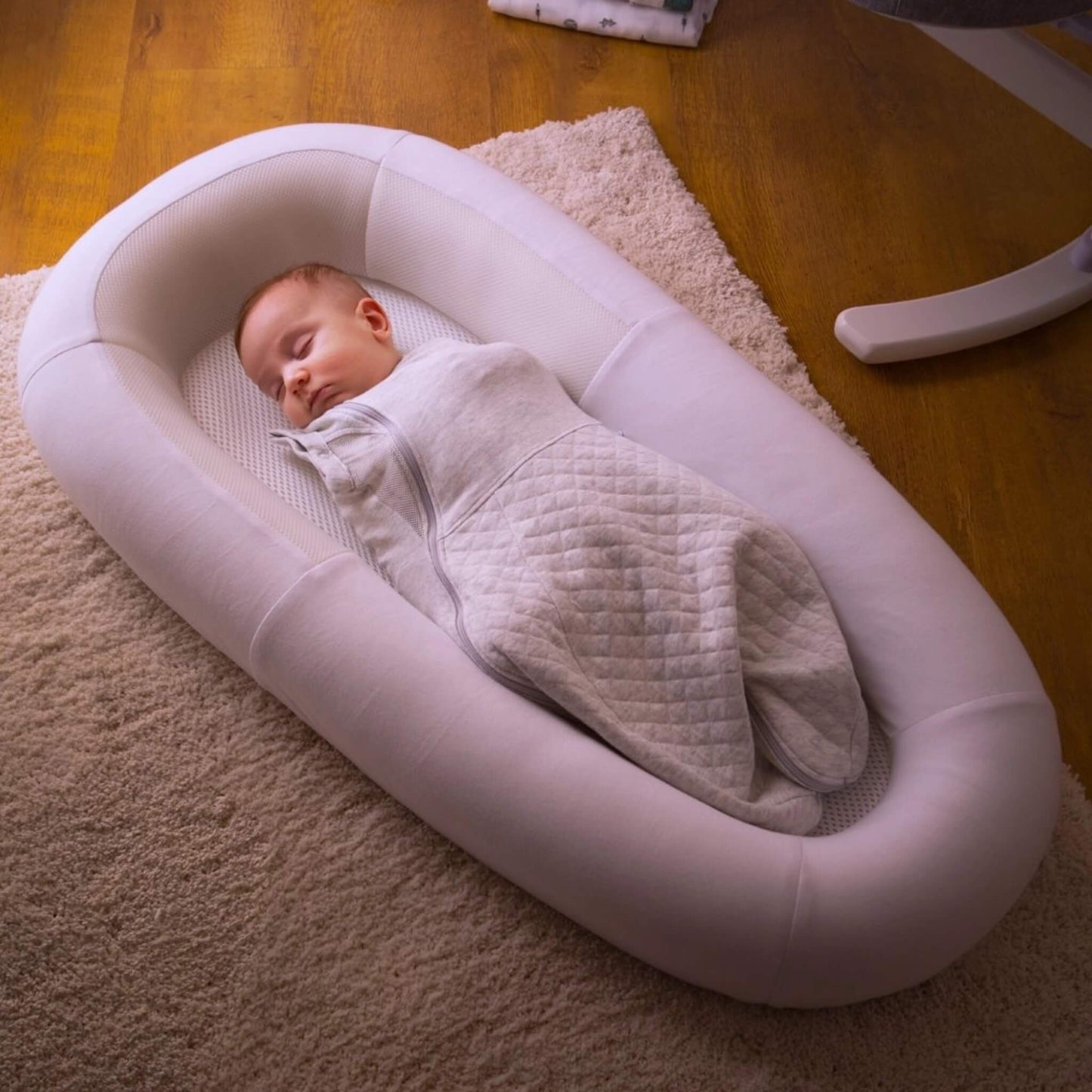 A baby sleeping while swaddled inside a structured baby bed with breathable mesh panels, positioned on a soft rug in a home environment.