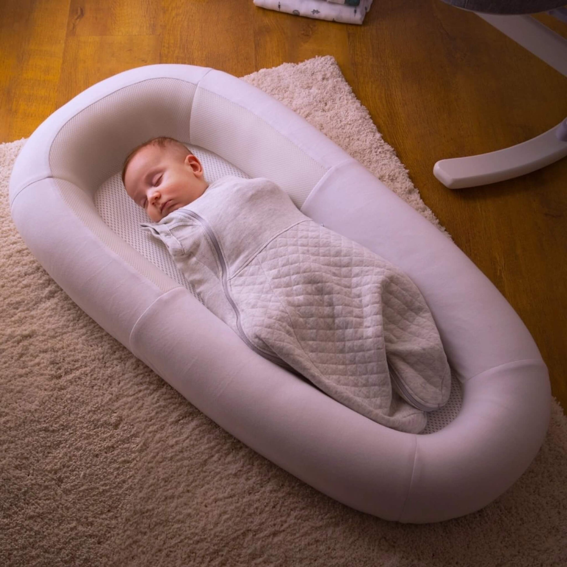 A baby sleeping while swaddled inside a structured baby bed with breathable mesh panels, positioned on a soft rug in a home environment.