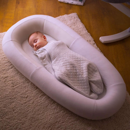 A baby sleeping while swaddled inside a structured baby bed with breathable mesh panels, positioned on a soft rug in a home environment.