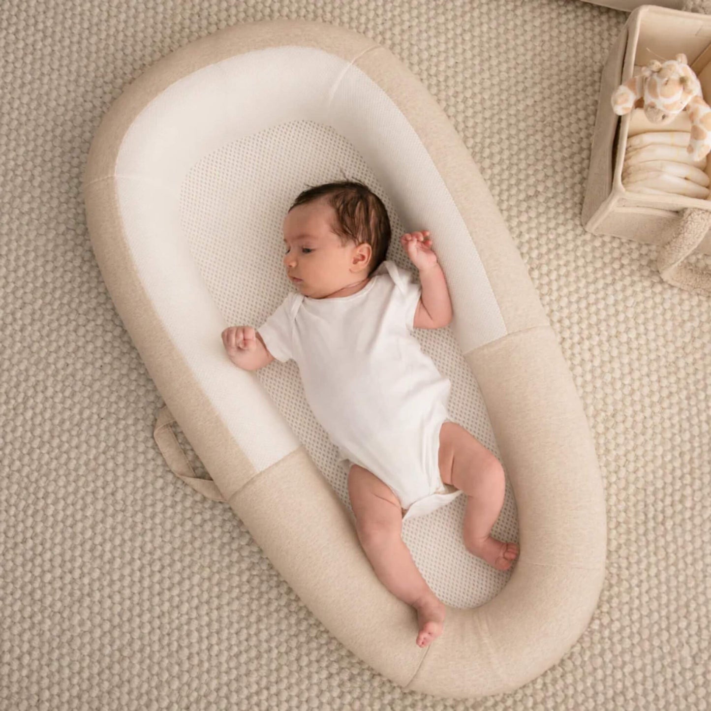 A baby lying on their back in a Sand coloured baby bed while wearing a white short-sleeved bodysuit.