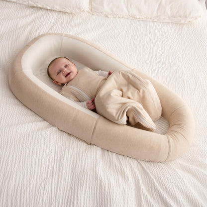 A smiling baby lying in a beige baby bed with a beige sleep bag draped over their legs.