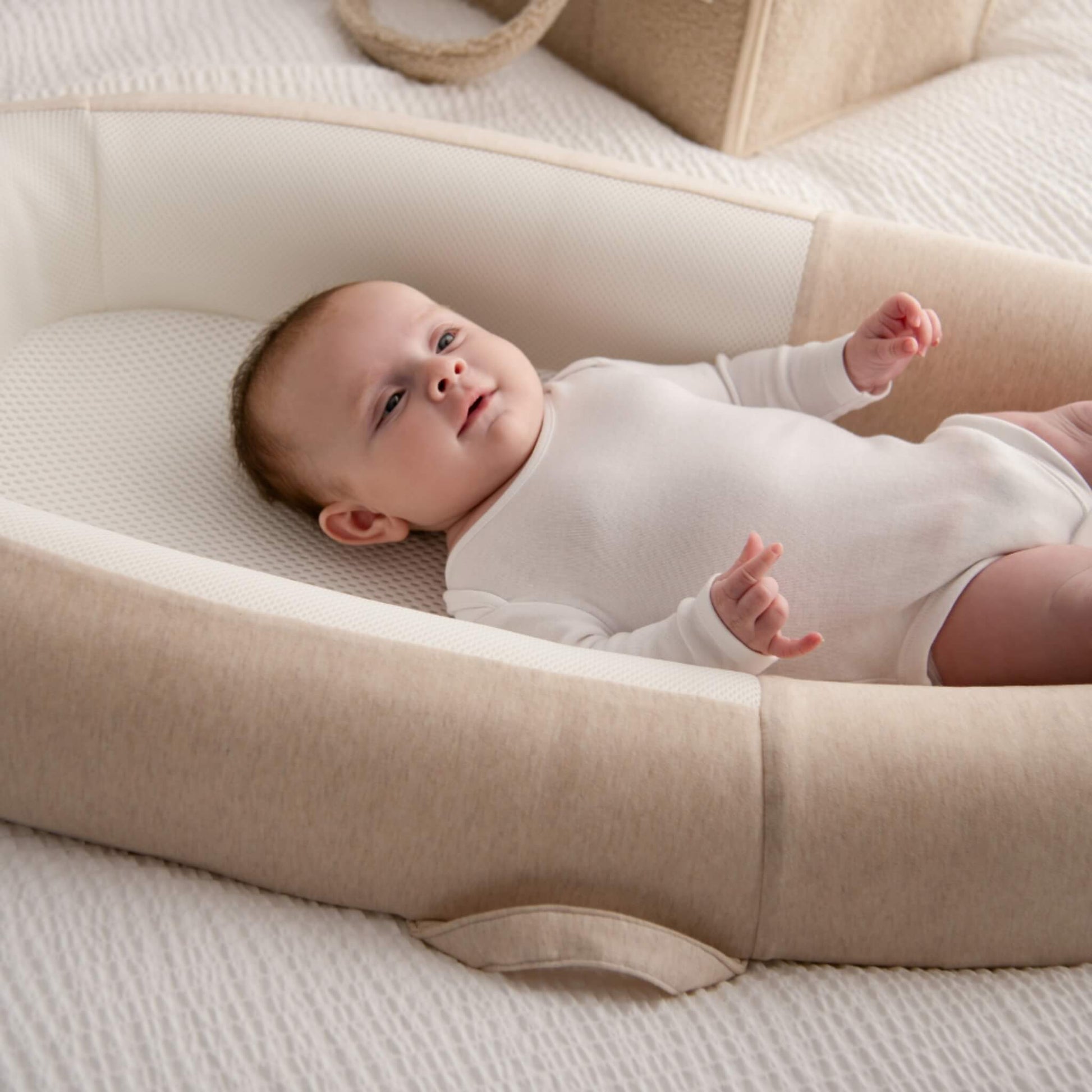 A smiling baby lying in a breathable mesh baby bed on a bed while wearing a white long-sleeved bodysuit.