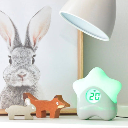 Star-shaped room thermometer glowing green on a nursery shelf beside wooden animal toys and a table lamp.