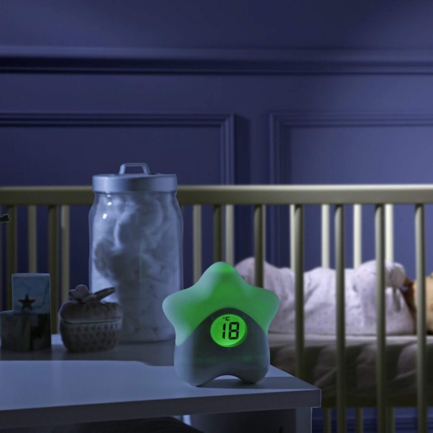 Star-shaped room thermometer glowing green on a bedside table next to a cot in a darkened nursery.