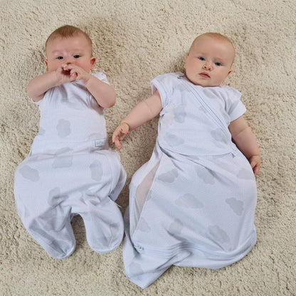 Two babies lying on a soft carpet wearing white with grey cloud-print swaddle sleep bags, showing arms in and arms out sleep stages side by side.