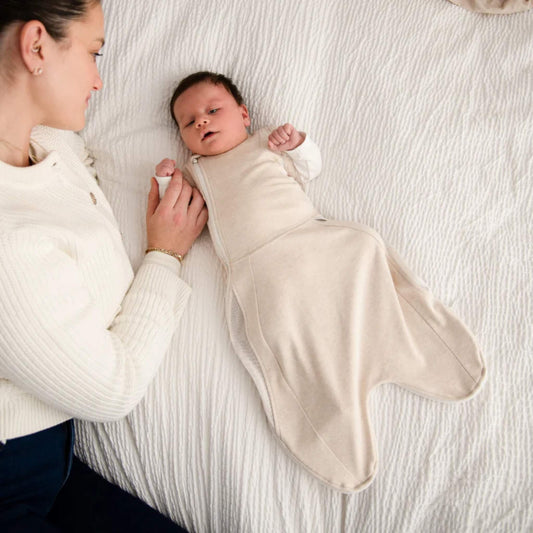 A baby wearing the Summer Sand swaddle to sleep bag while lying on a bed beside an adult holding their hand.